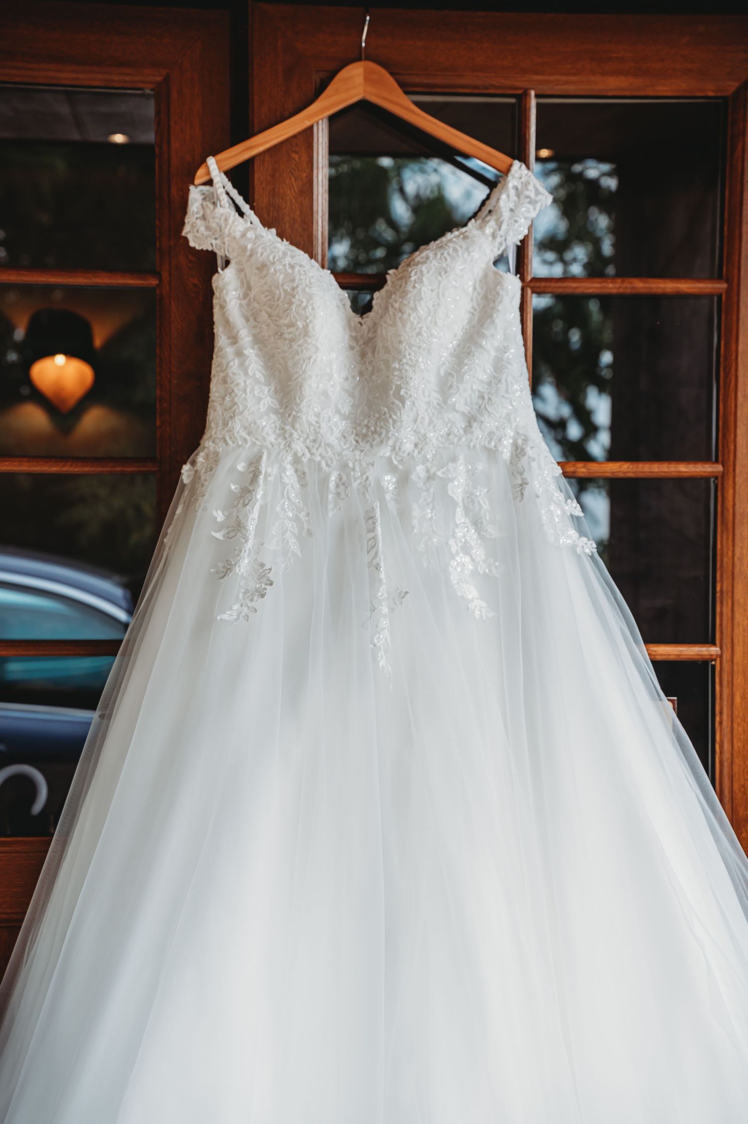 Elegant white wedding gown with lace detailed bodice and tulle skirt hangs on wooden hanger against window.
