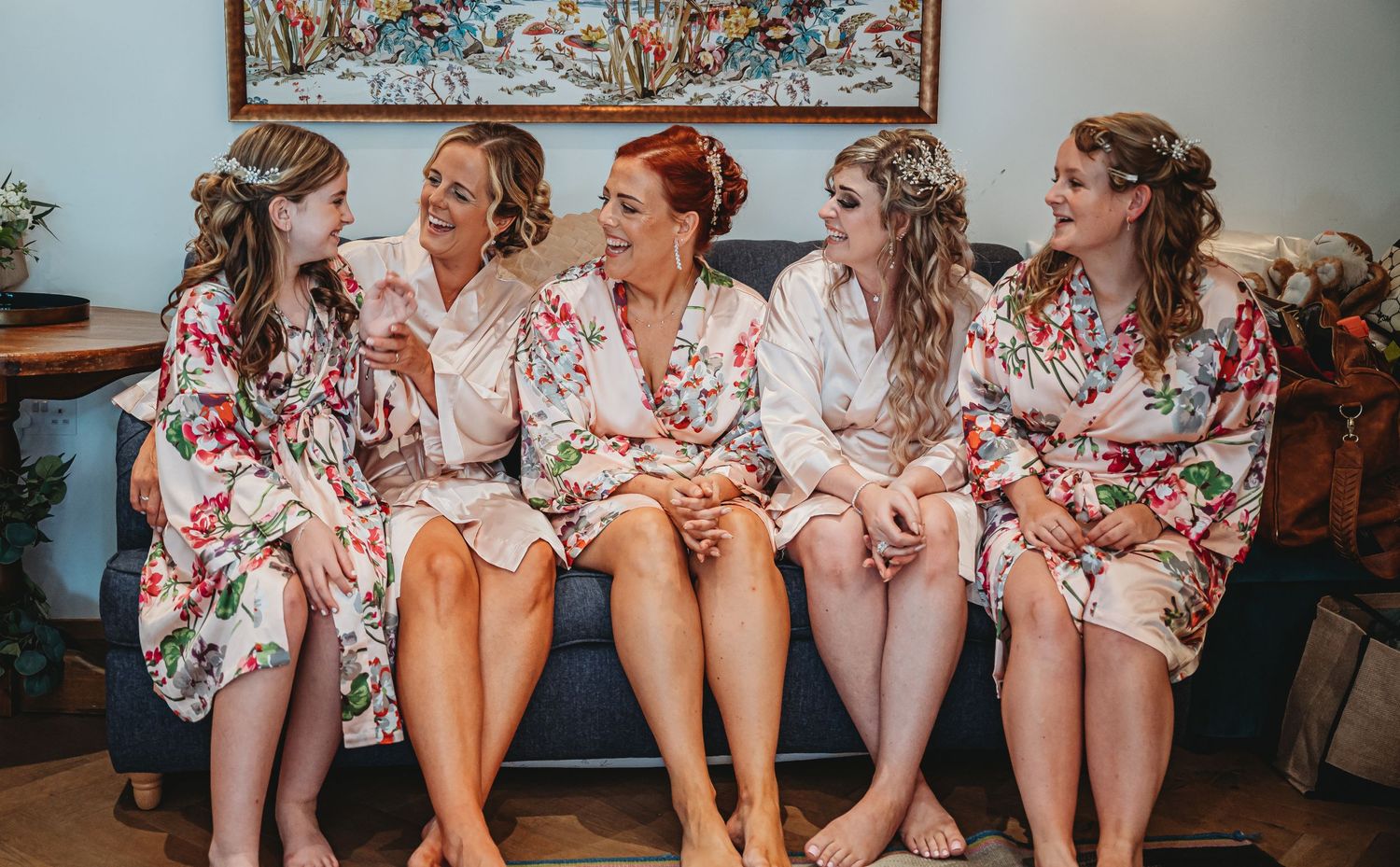 A group of five people wearing matching floral robes sit together on a couch during a wedding preparation.