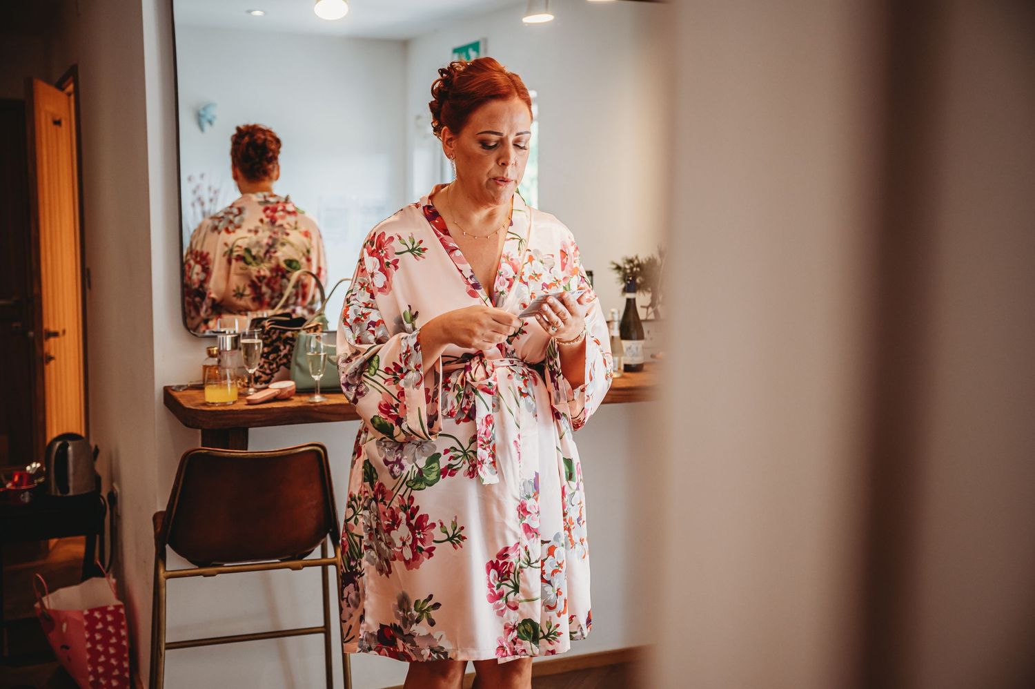 Someone in a pink floral robe with red hair stands in a room looking at their phone before a dressing room mirror.