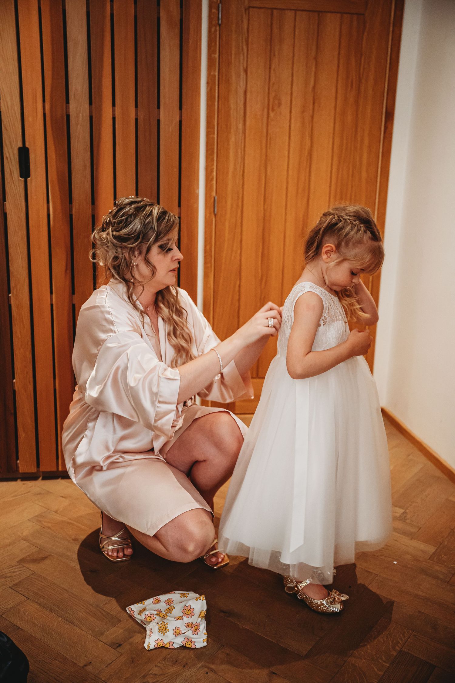 A touching wedding preparation moment with a woman helping a young girl in a white flower girl dress on hardwood floors.
