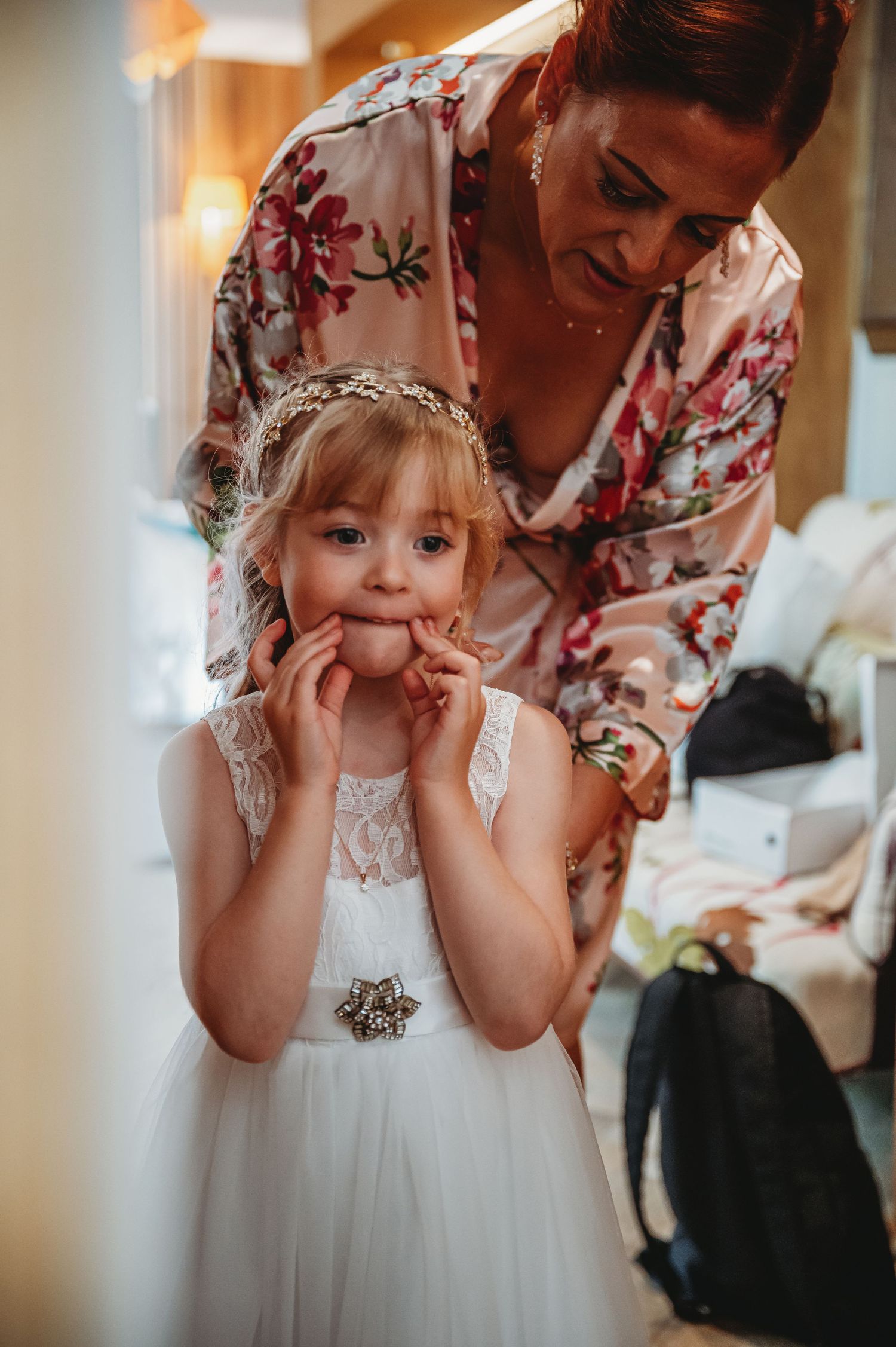 A young flower girl in a white lace dress and headpiece stands nervously while being helped with preparations.