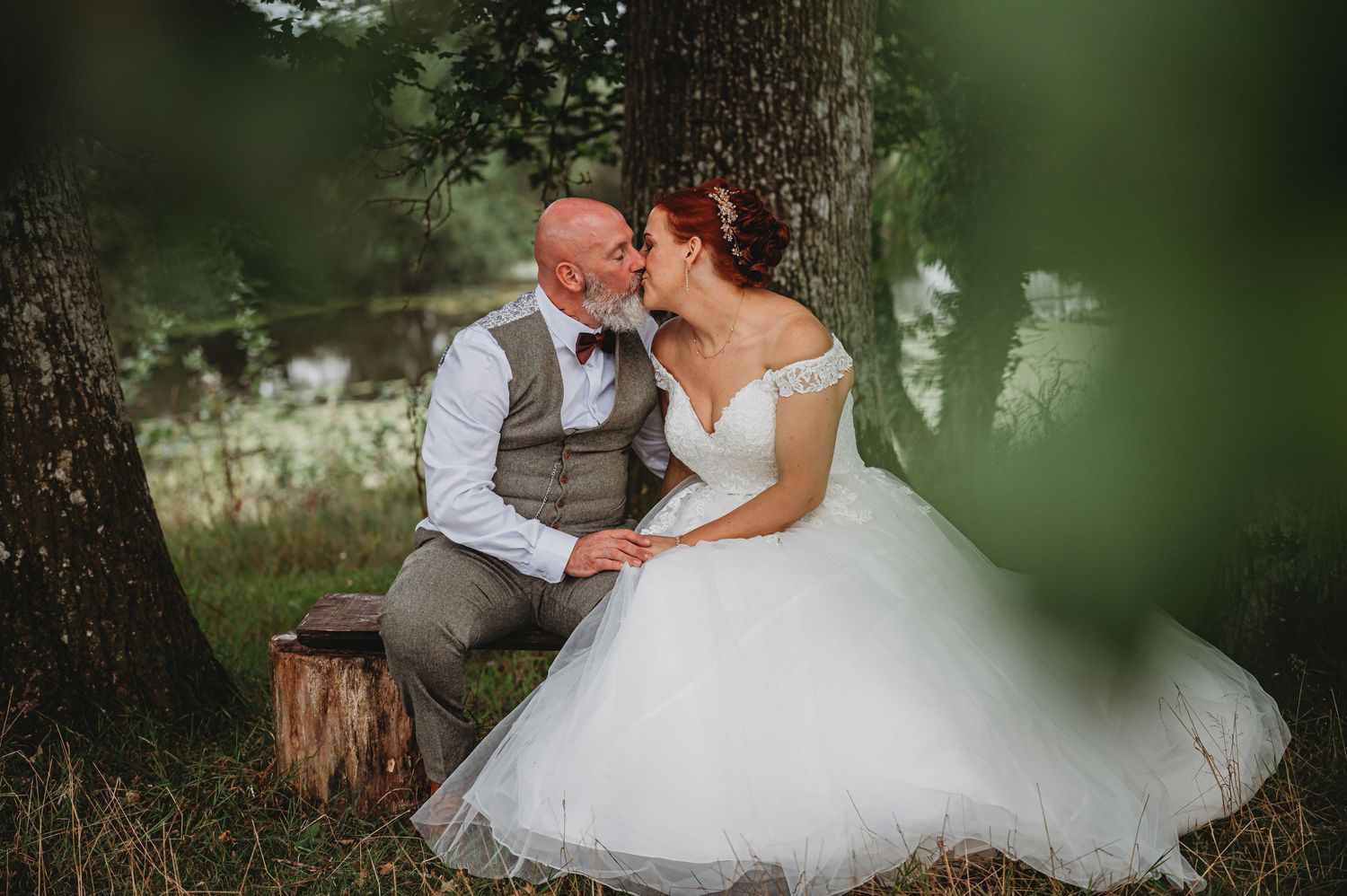A couple shares a romantic moment outdoors on their wedding day, seated by a tree in formal attire.