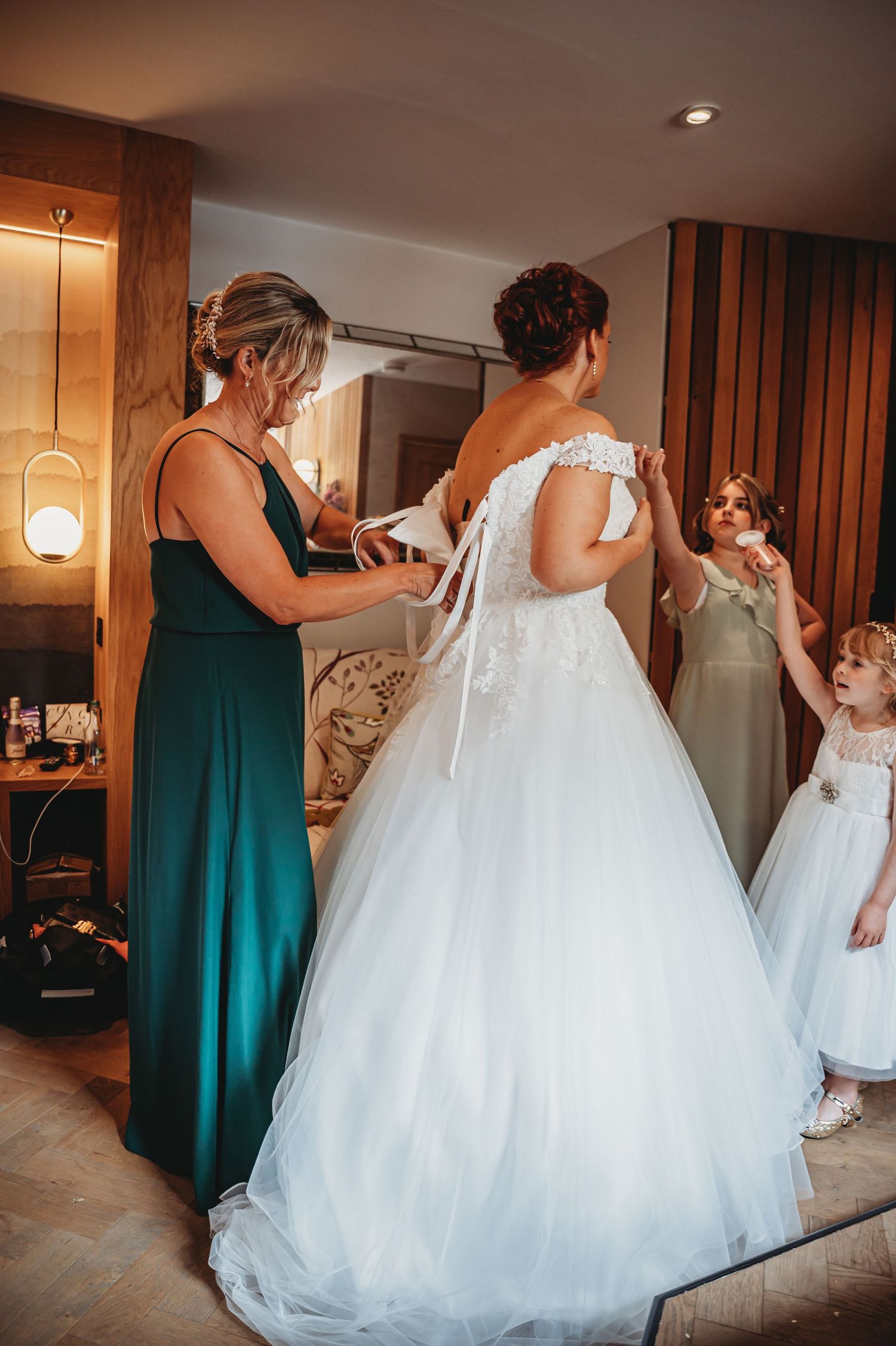 A bride in a white lace ballgown gets help fastening her dress while wearing an elegant off-shoulder wedding gown.