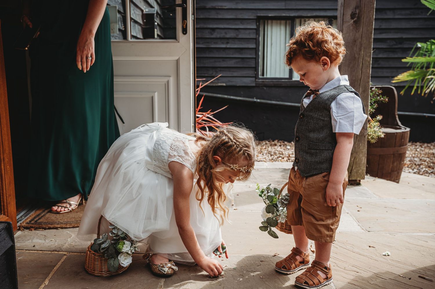 Two children in vintage-style clothing explore together on a porch with brown suspenders and a white dress.