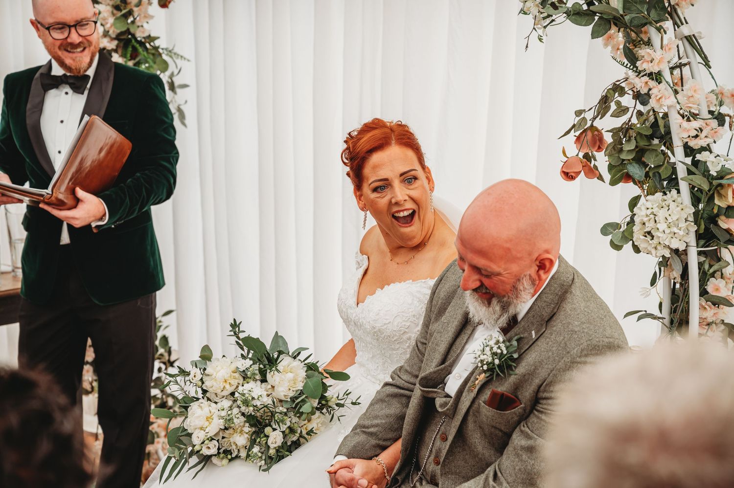 A candid moment of laughter during a wedding ceremony with white floral decorations and a violinist in the background.