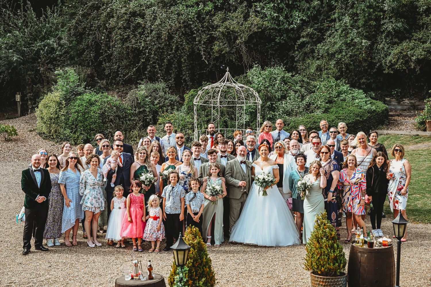 Large wedding party group gathered outdoors for formal photo in front of decorative garden arbor.