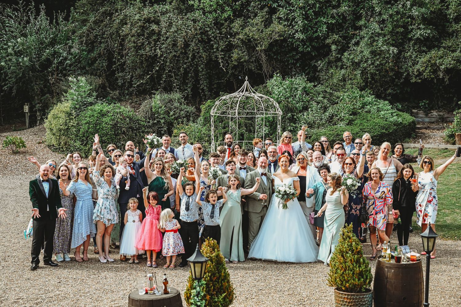 A large group of wedding guests pose with formal attire in front of a garden archway surrounded by greenery.