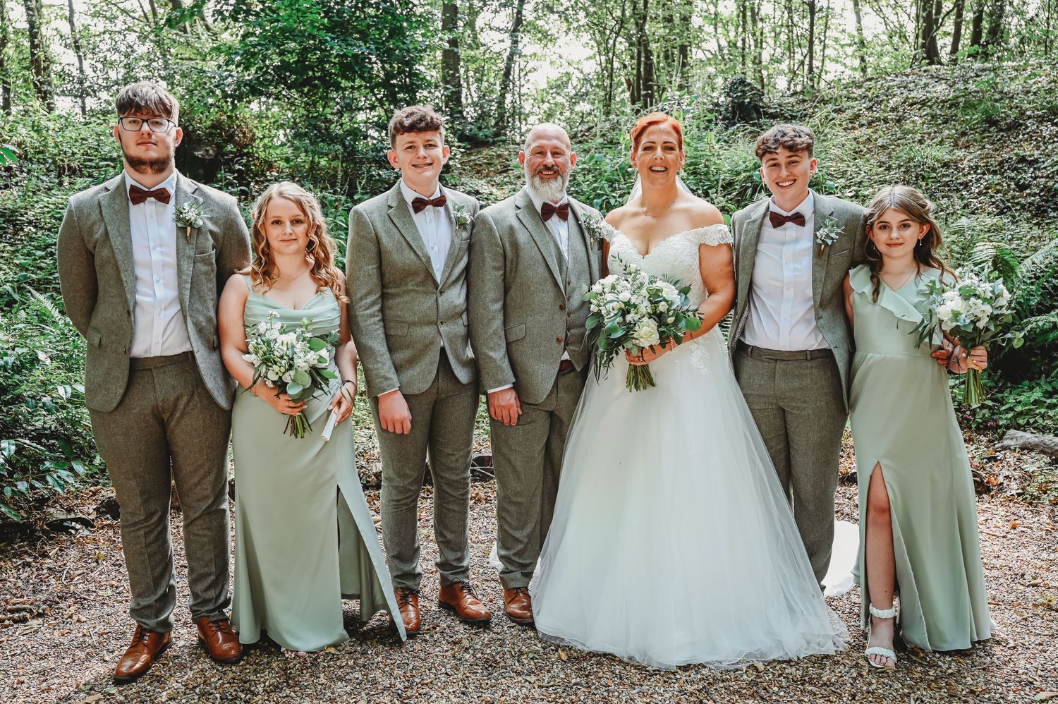 Wedding party in sage green and gray attire poses together in a wooded forest setting holding white floral bouquets.