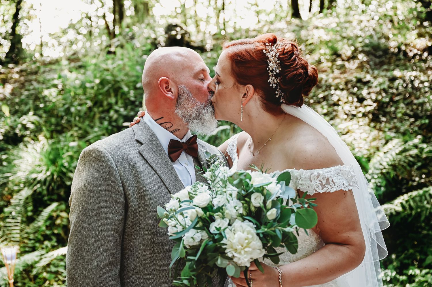 A couple shares an intimate moment in a sunlit forest while holding a green and white wedding bouquet.