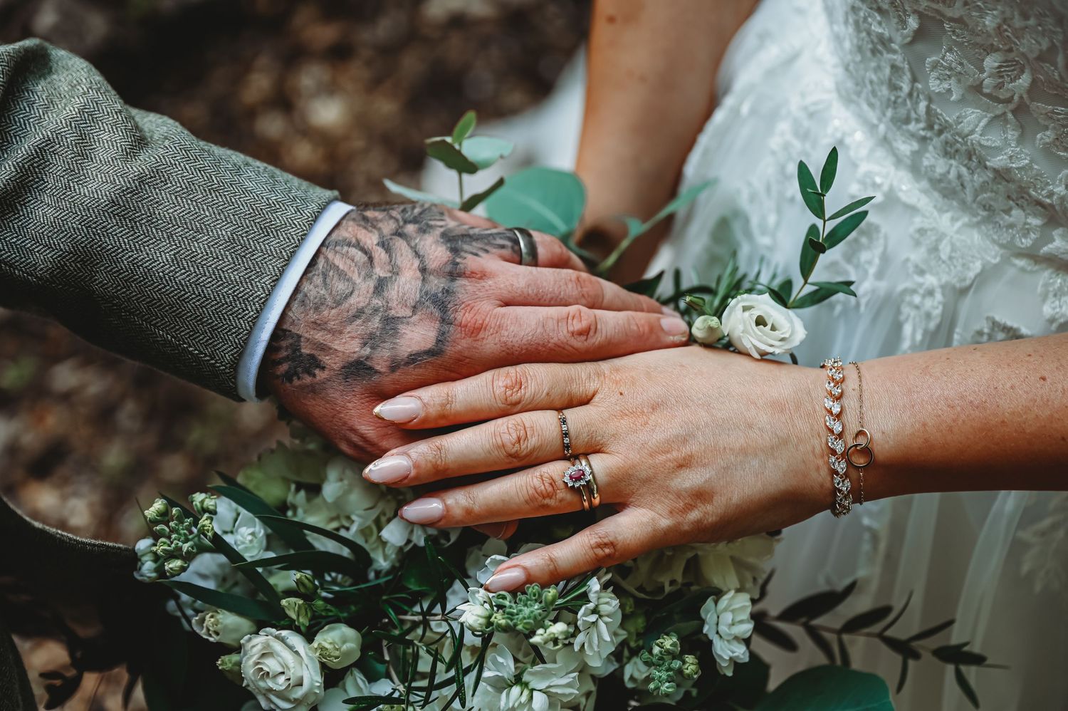 Two hands wearing wedding rings rest together on a bouquet of white flowers and greenery.