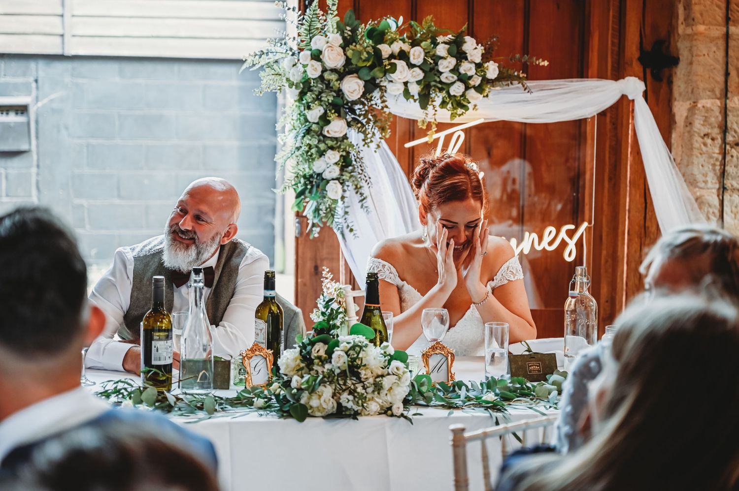 Emotional reaction during a wedding reception speech at a rustic venue decorated with eucalyptus and white floral arrangements.