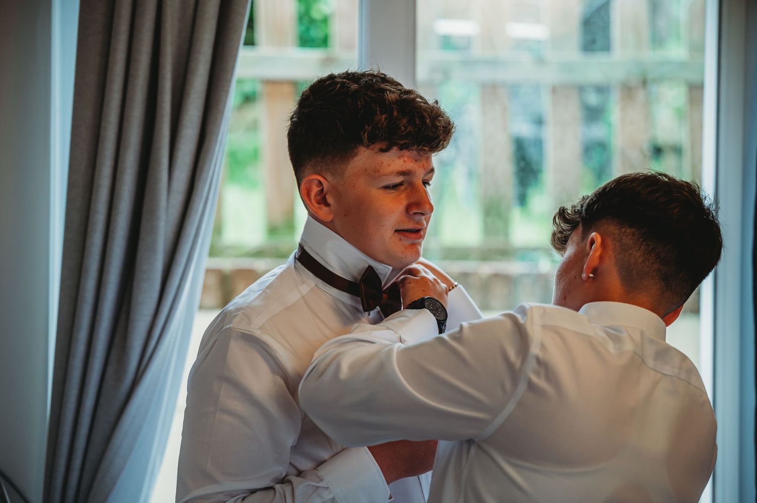 Two people in white dress shirts help each other with bow ties by a window with curtains and natural light.