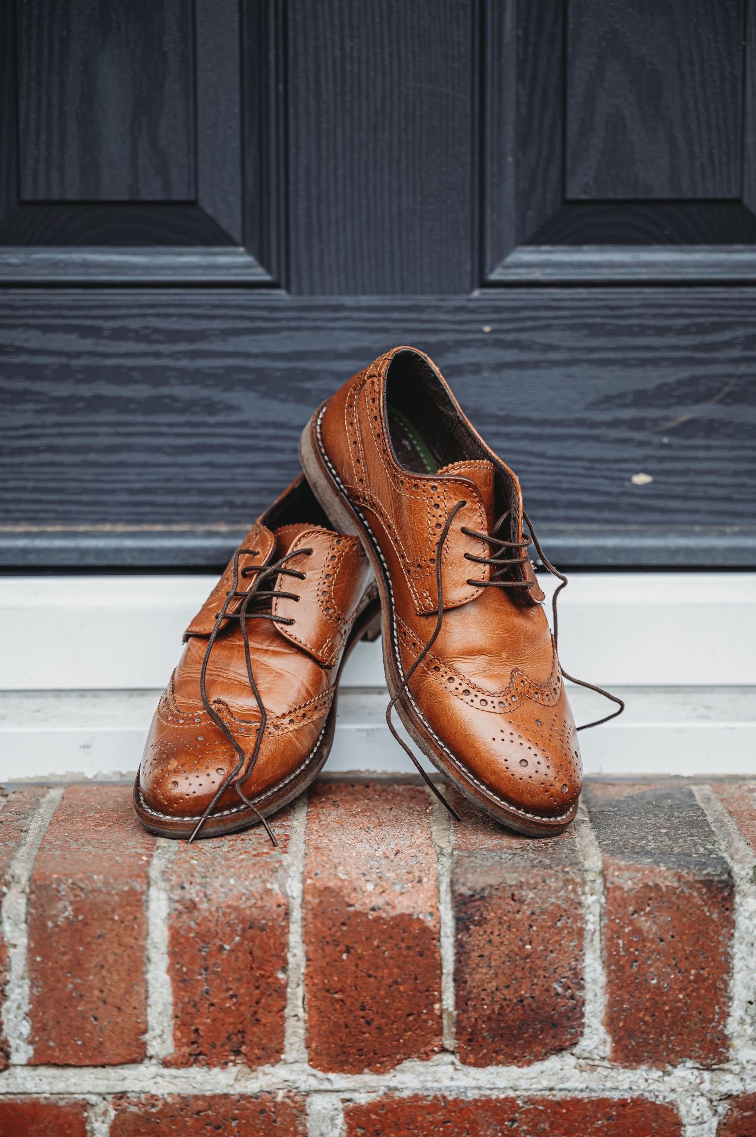 A pair of classic brown leather wingtip oxford dress shoes placed on red brick steps against a dark door.