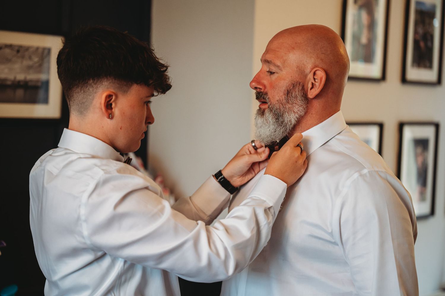 Two people share a tender moment while adjusting a white dress shirt collar in a room with framed photos on the wall.