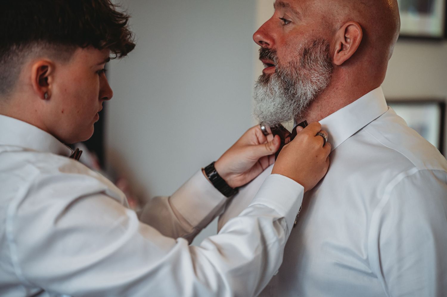 Two people stand together dressed in white dress shirts while one helps adjust the other's necktie.