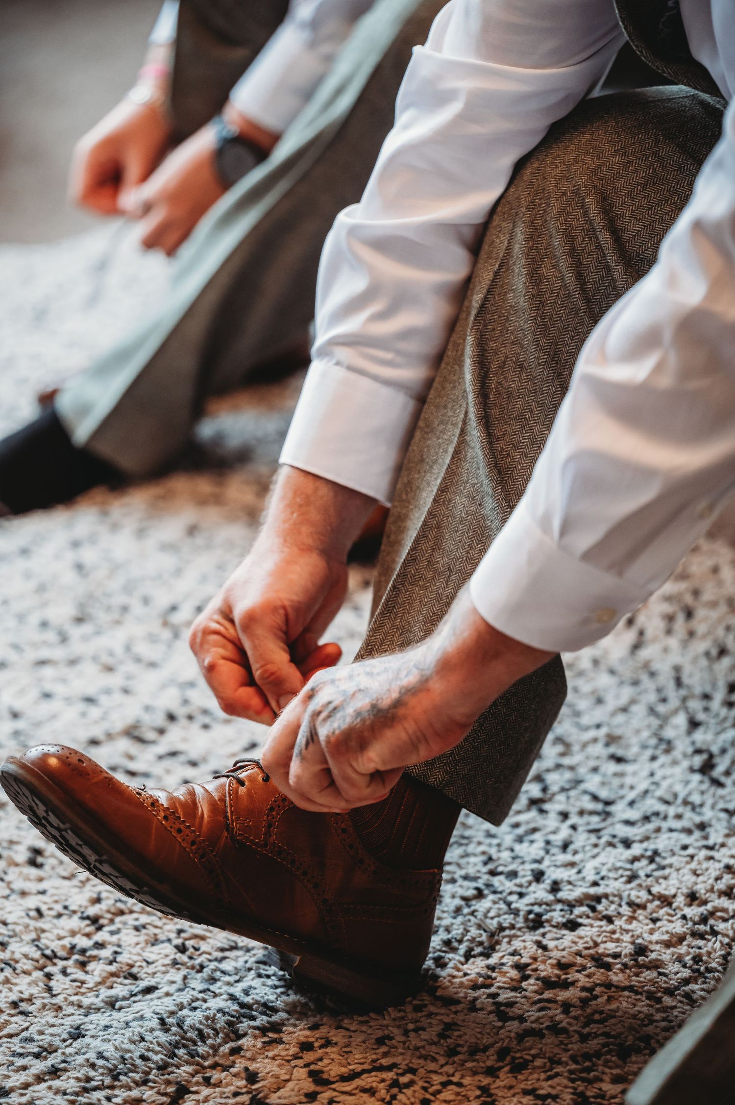 Person tying brown leather dress shoes while wearing a white shirt and grey pants on a textured carpet.