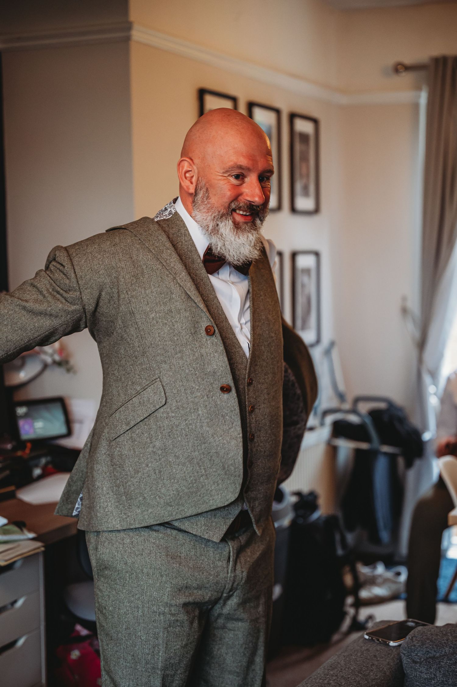 A bearded gentleman in a grey tweed suit smiles while standing in a home office setting.