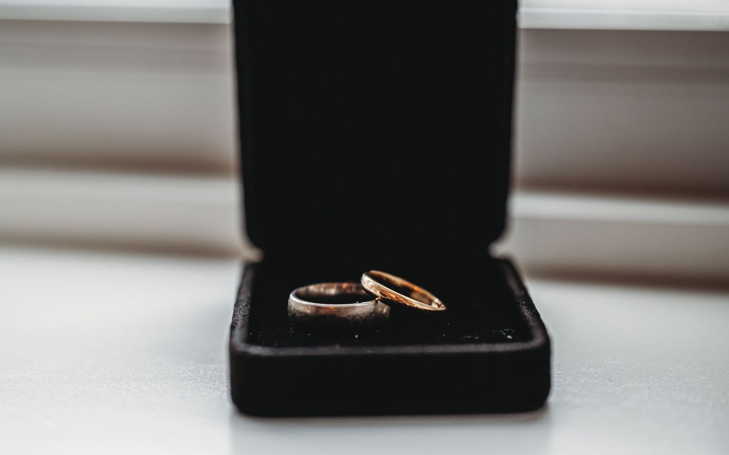Two gold wedding rings rest in an open black jewelry box near a window with soft natural lighting.