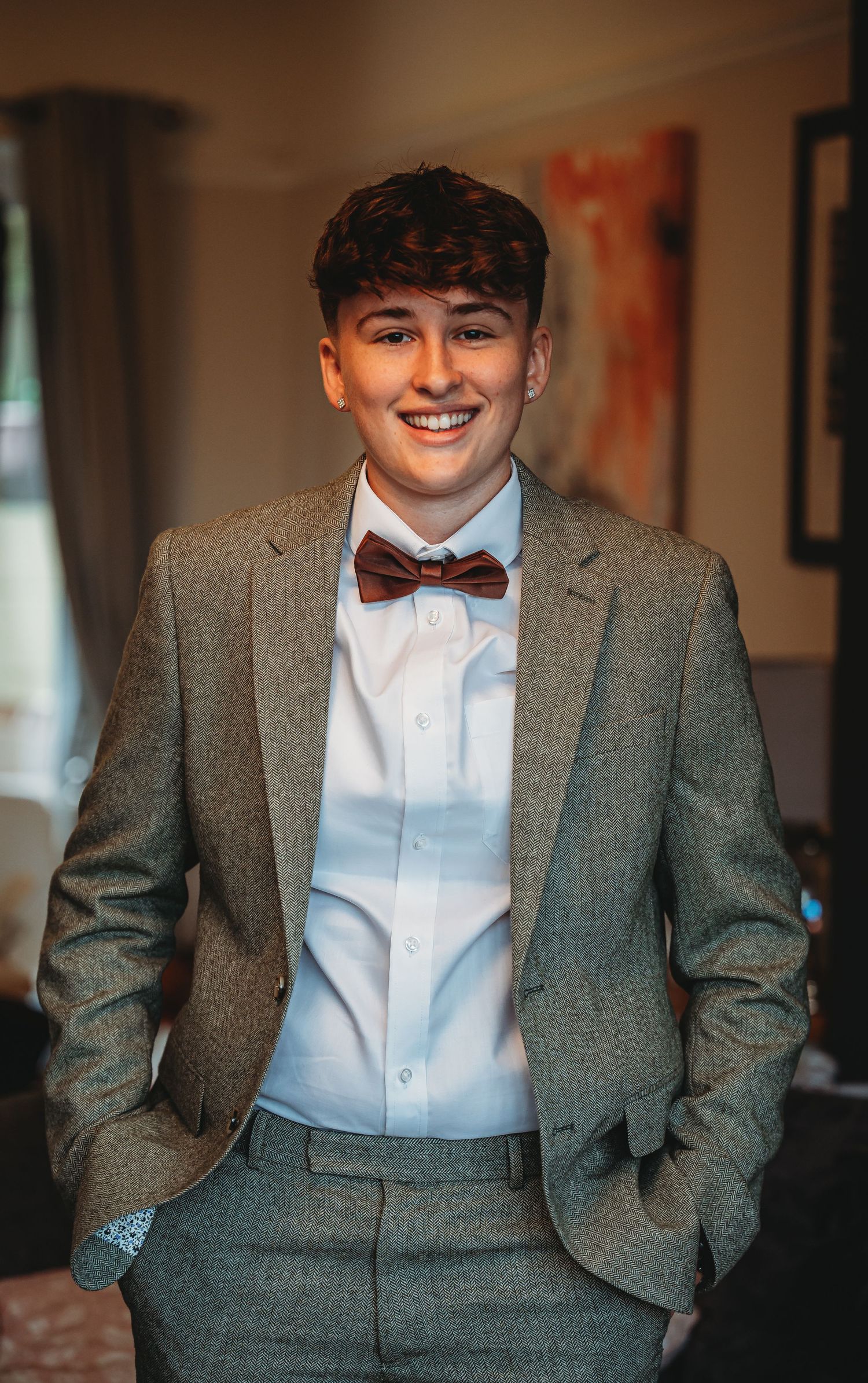 Young man in grey suit and burgundy bow tie poses confidently in a room with artwork.