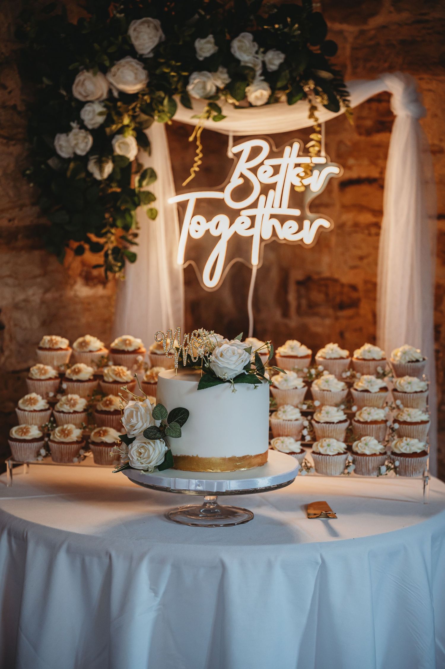 Wedding dessert table with blue and gold cake, cupcakes and 'Better Together' neon sign under floral arch decor.