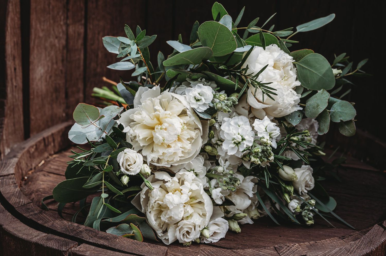 A romantic white peony and garden rose bouquet with eucalyptus greenery rests on rustic wooden surface.