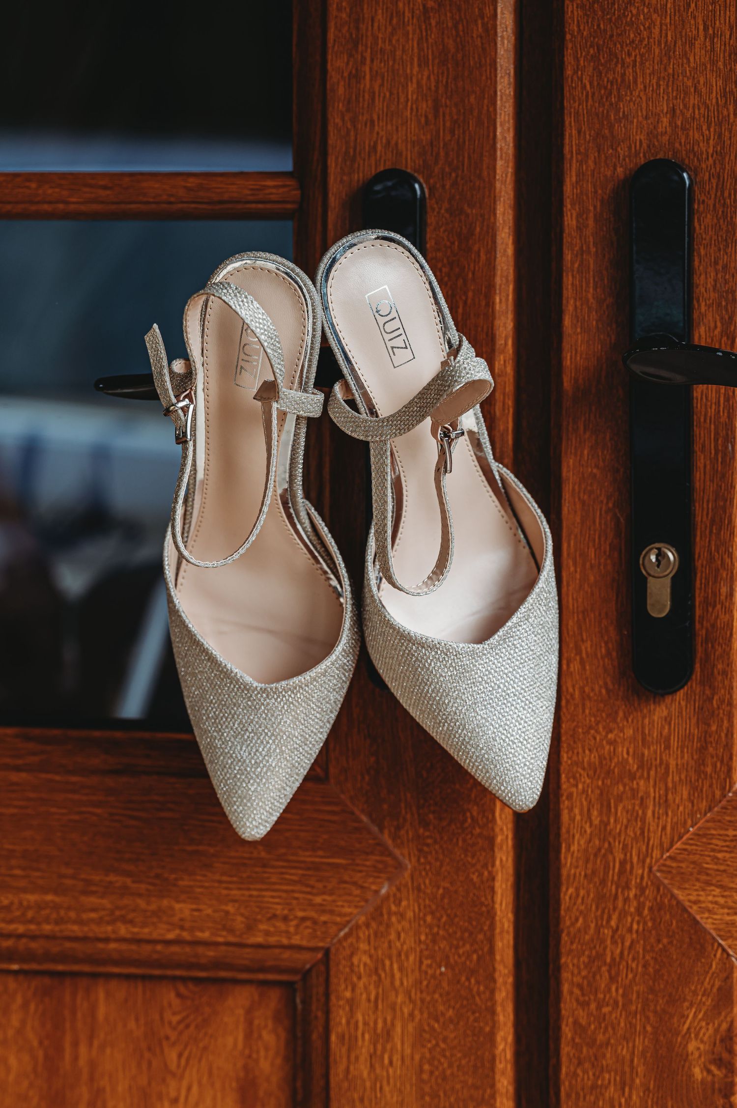 A pair of sparkly champagne-colored pointed toe slingback heels hanging on a wooden door handle.