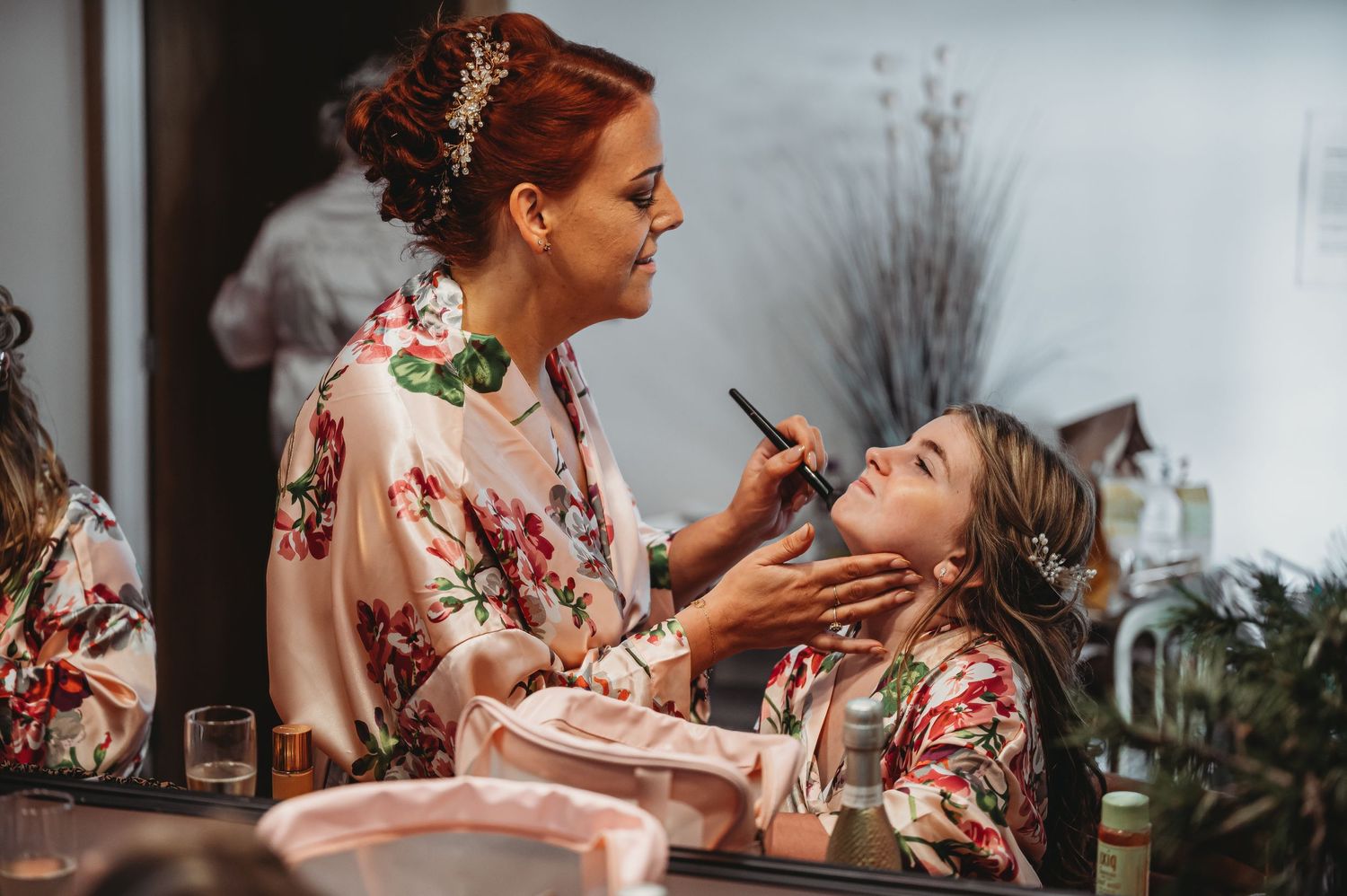 A makeup artist applying cosmetics on someone wearing a matching floral robe during wedding preparation.