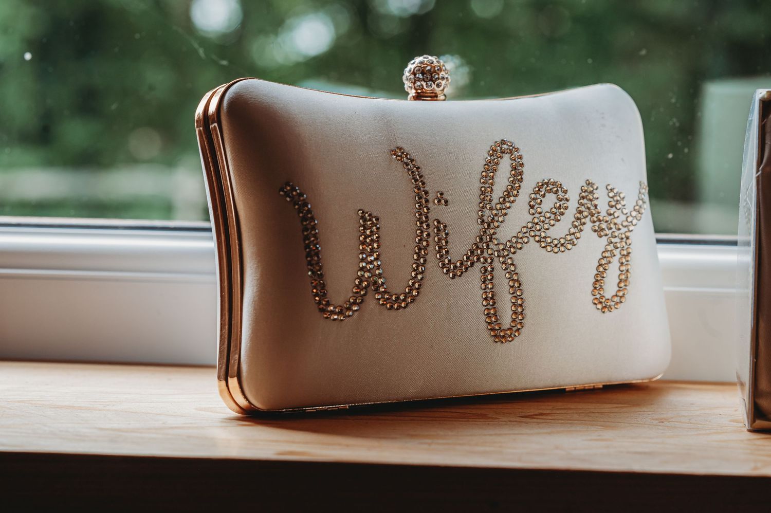 White bridal clutch purse with 'Wifey' written in rhinestone detail sits on wooden windowsill.