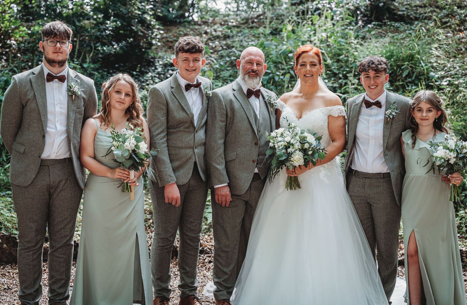 Wedding party in sage green dresses and gray suits pose together in a lush outdoor garden setting.