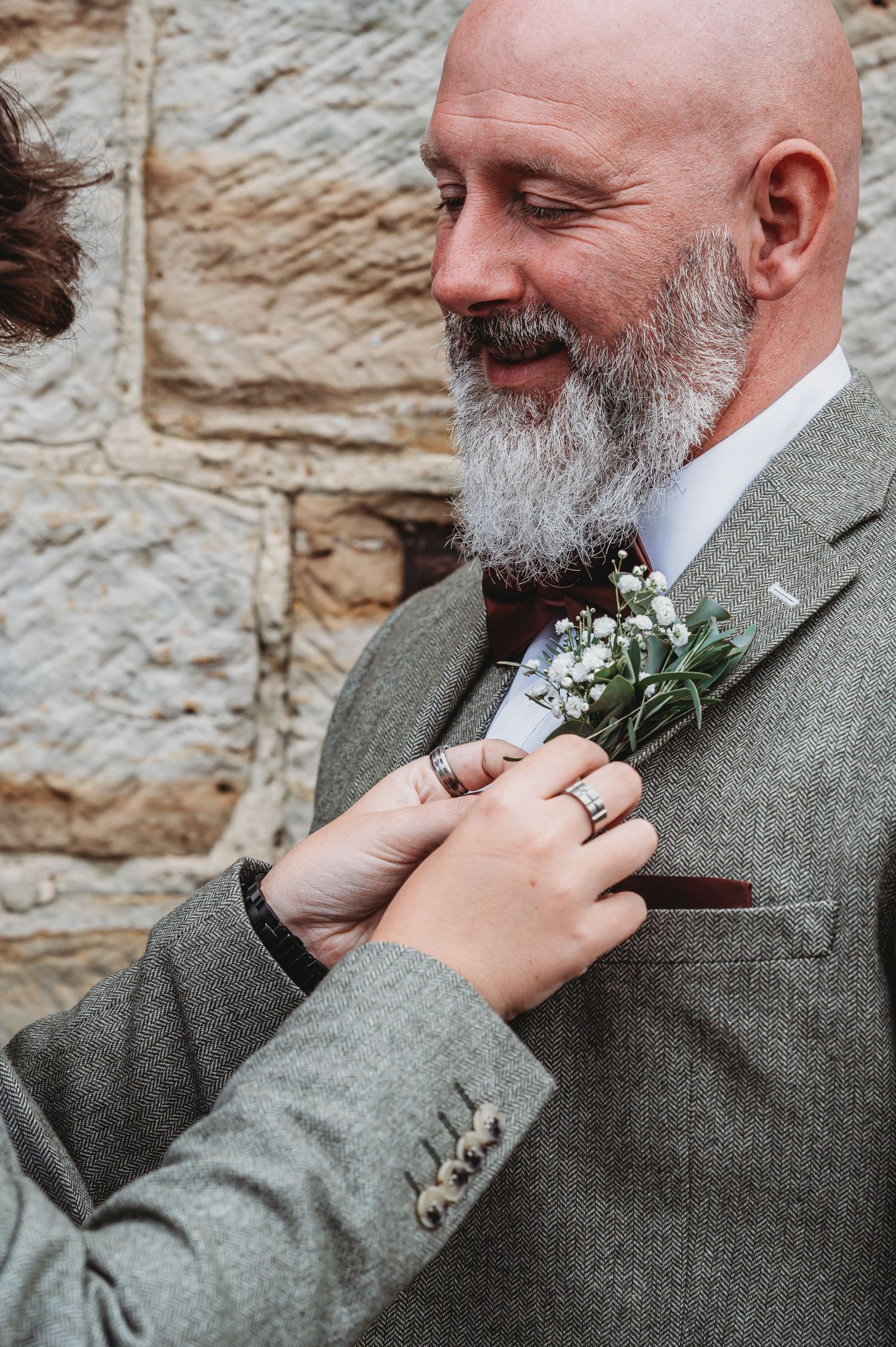 Hands pin delicate white floral boutonniere onto grey suit lapel during wedding preparation.