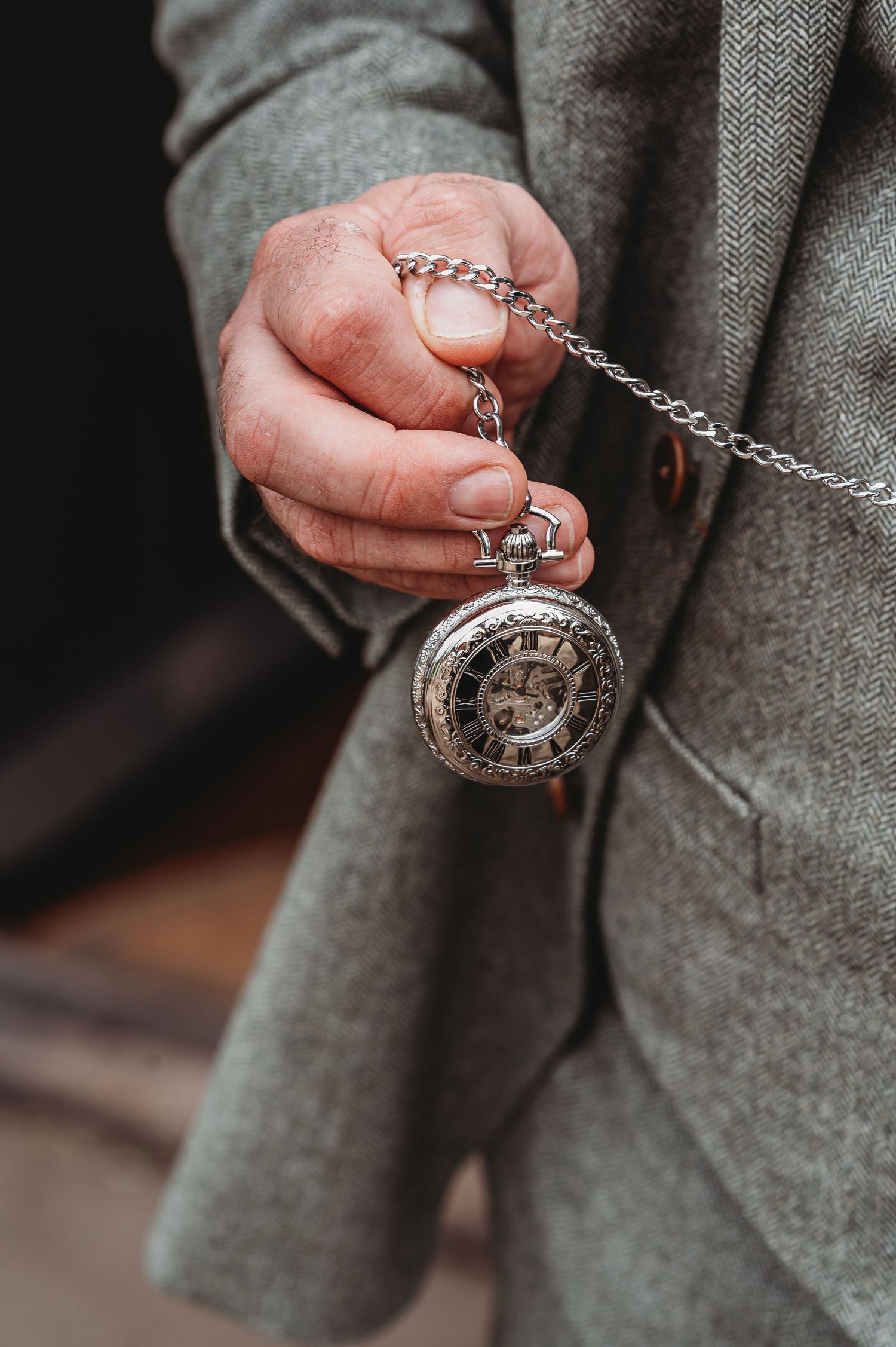 Hand holding vintage silver pocket watch with chain against grey coat sleeve in moody lighting.