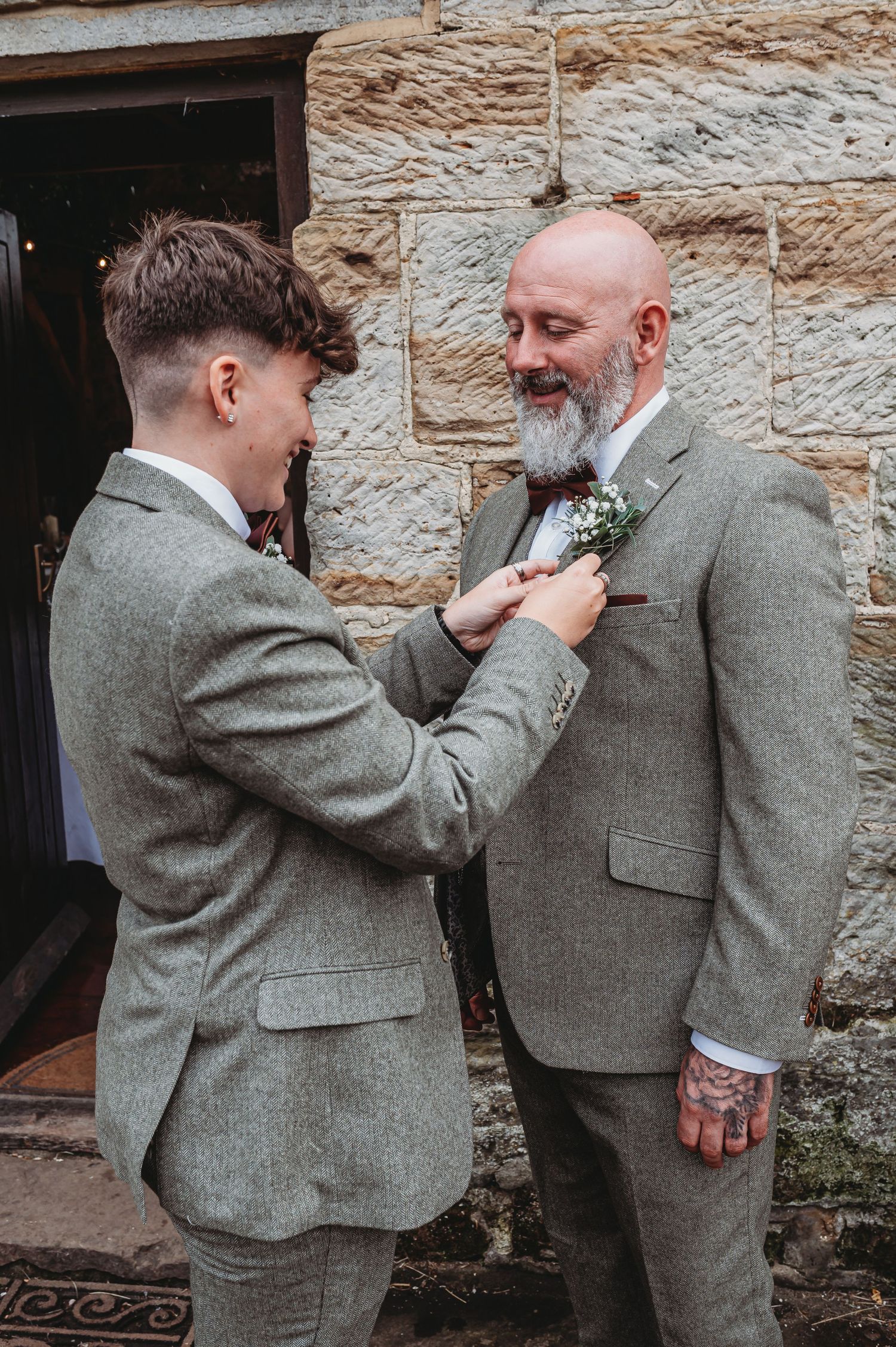 Two people in matching gray tweed suits share a formal moment as one adjusts the other's boutonniere against a stone wall.