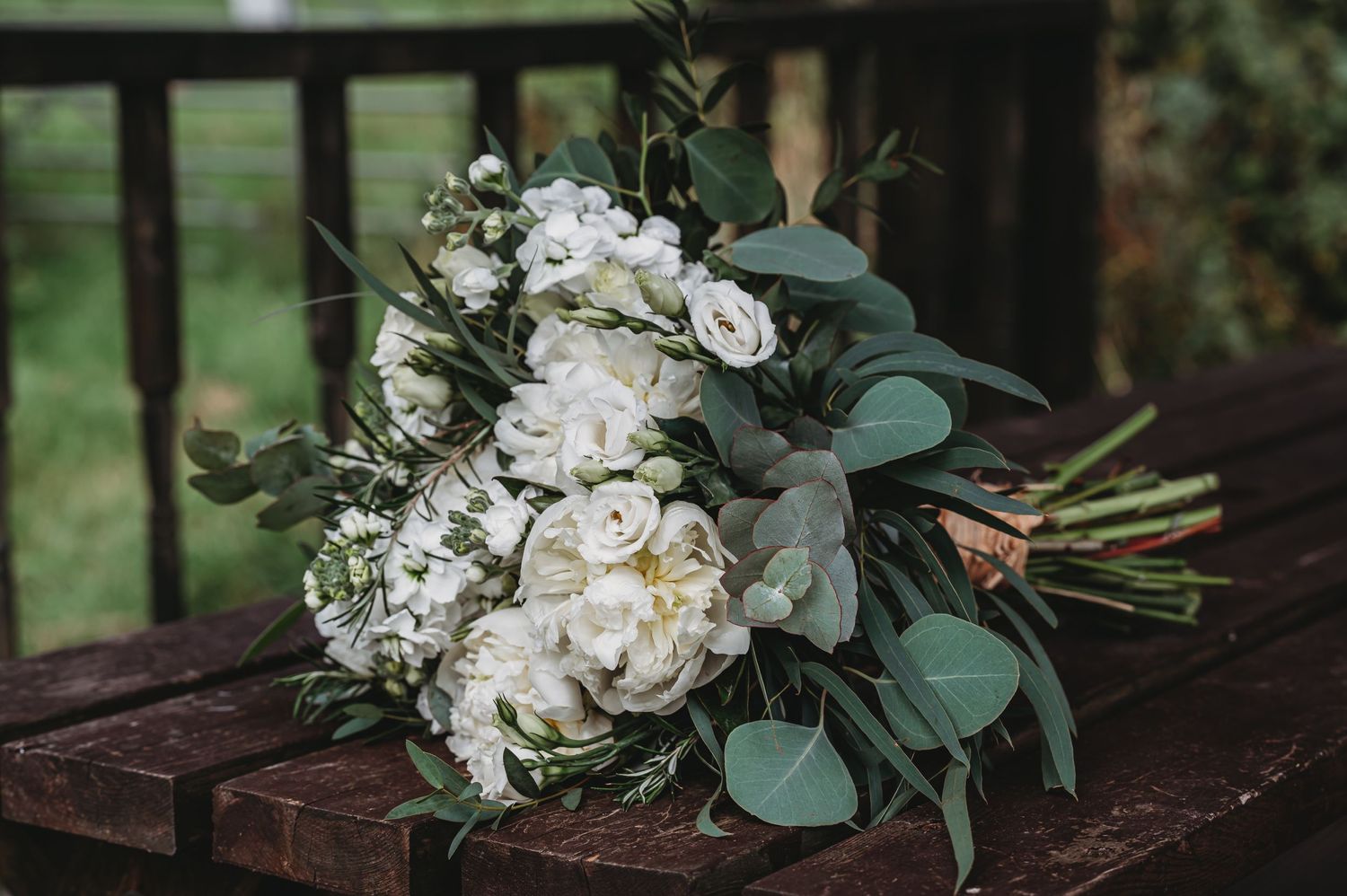 A stunning white bridal bouquet with roses and eucalyptus leaves rests on a dark wooden bench.