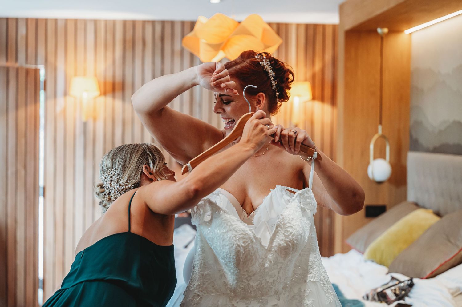 Person in emerald green dress helps someone put on a white lace wedding gown in a wood-paneled room.