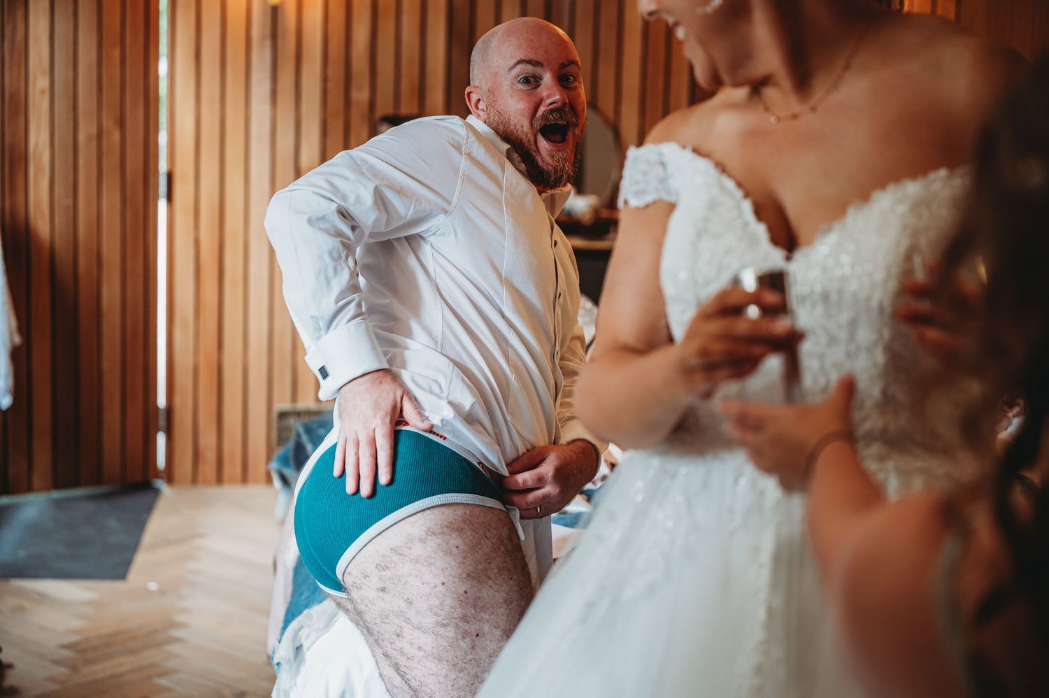 A person in swim trunks poses dramatically next to someone wearing a white wedding dress in a wood-paneled room.