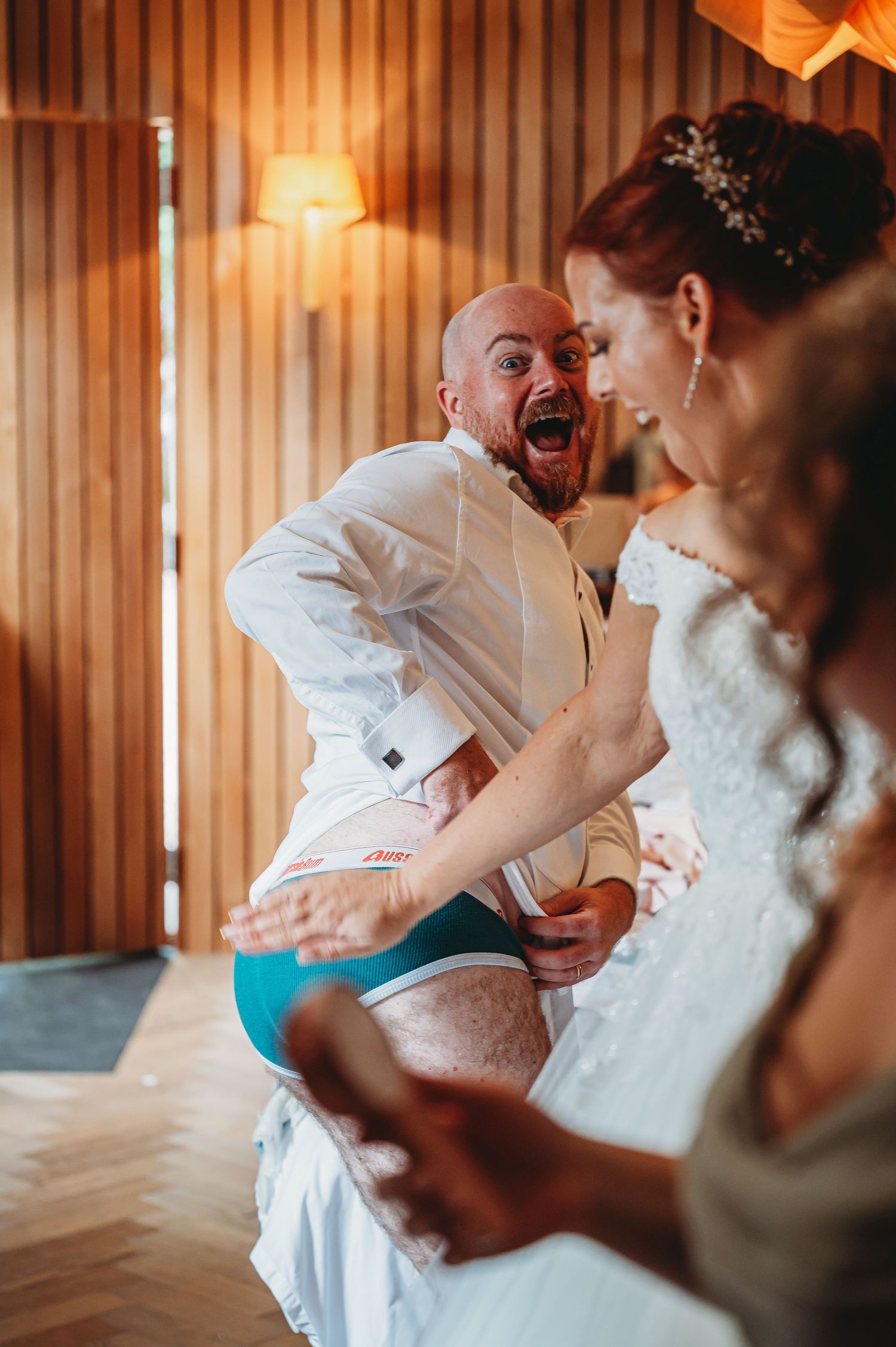 A groom in a white dress shirt and turquoise shorts shows excitement while preparing for his wedding ceremony.