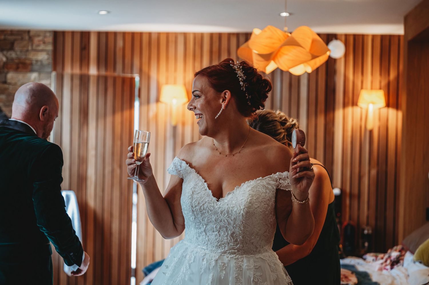 A person in a white off-shoulder wedding dress laughs while holding a glass during an indoor reception.