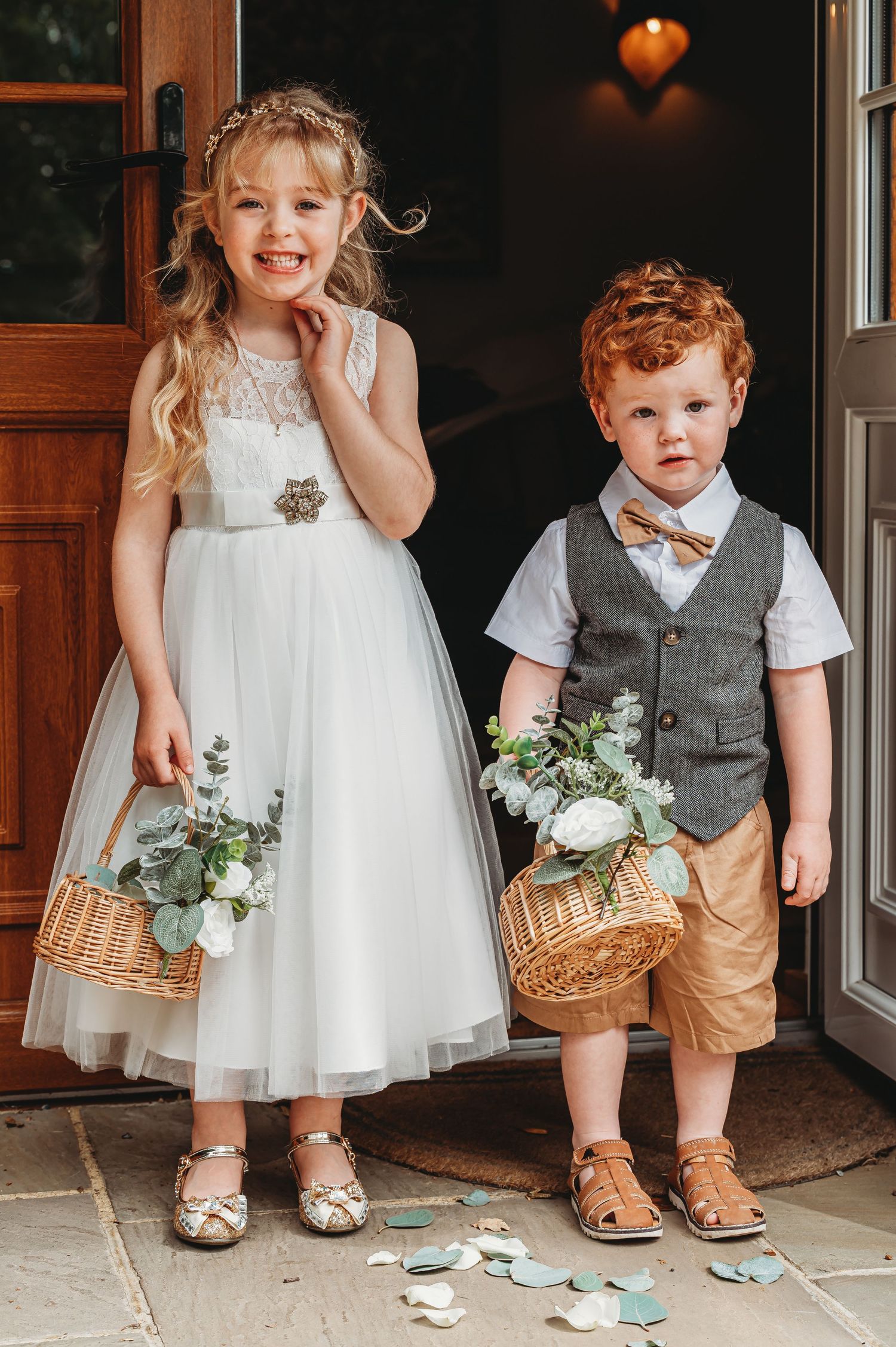 Two young children in wedding attire hold wicker baskets with flowers standing in a doorway.