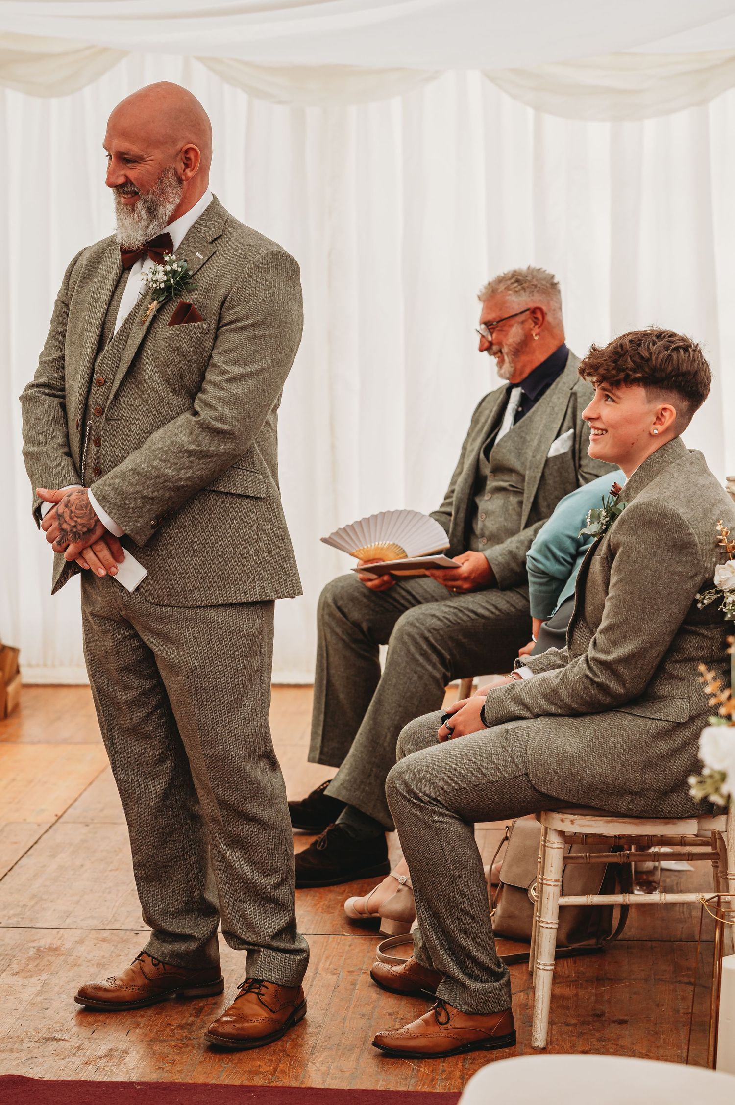 Men in grey suits standing and seated at a wedding ceremony with white draped backdrop and wooden chairs.