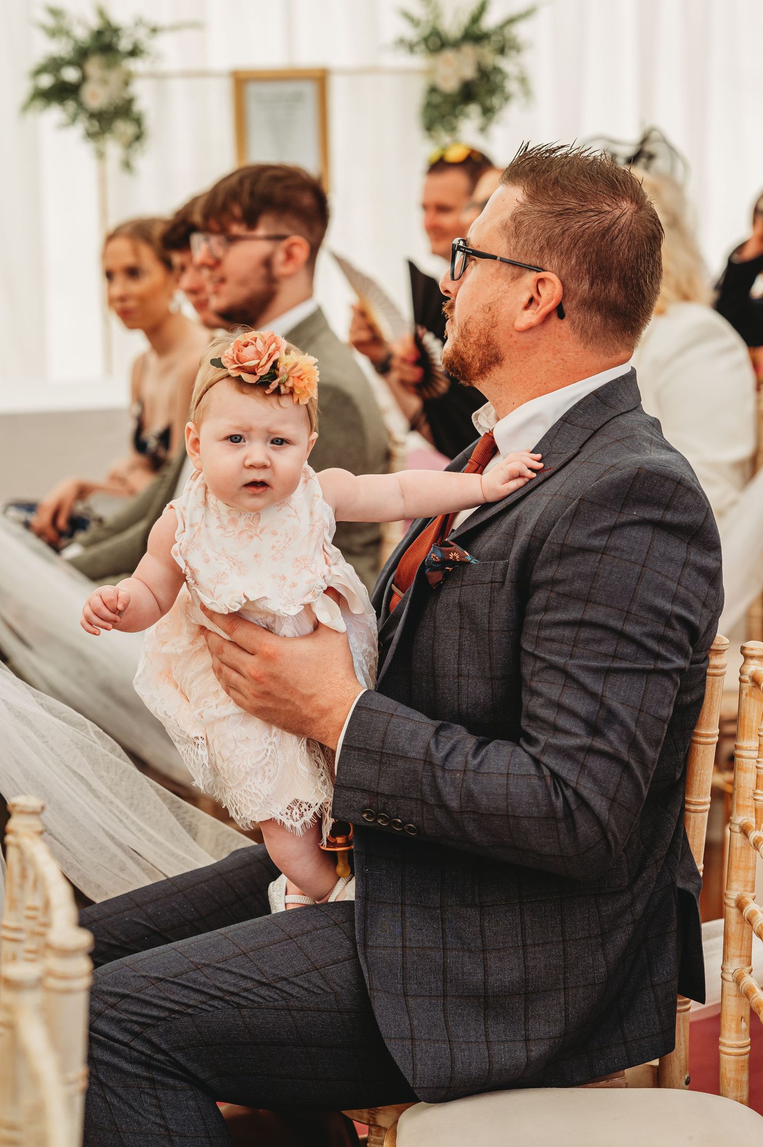 A baby wearing a white dress and floral crown sits on the lap of a guest in a gray suit during a wedding ceremony.