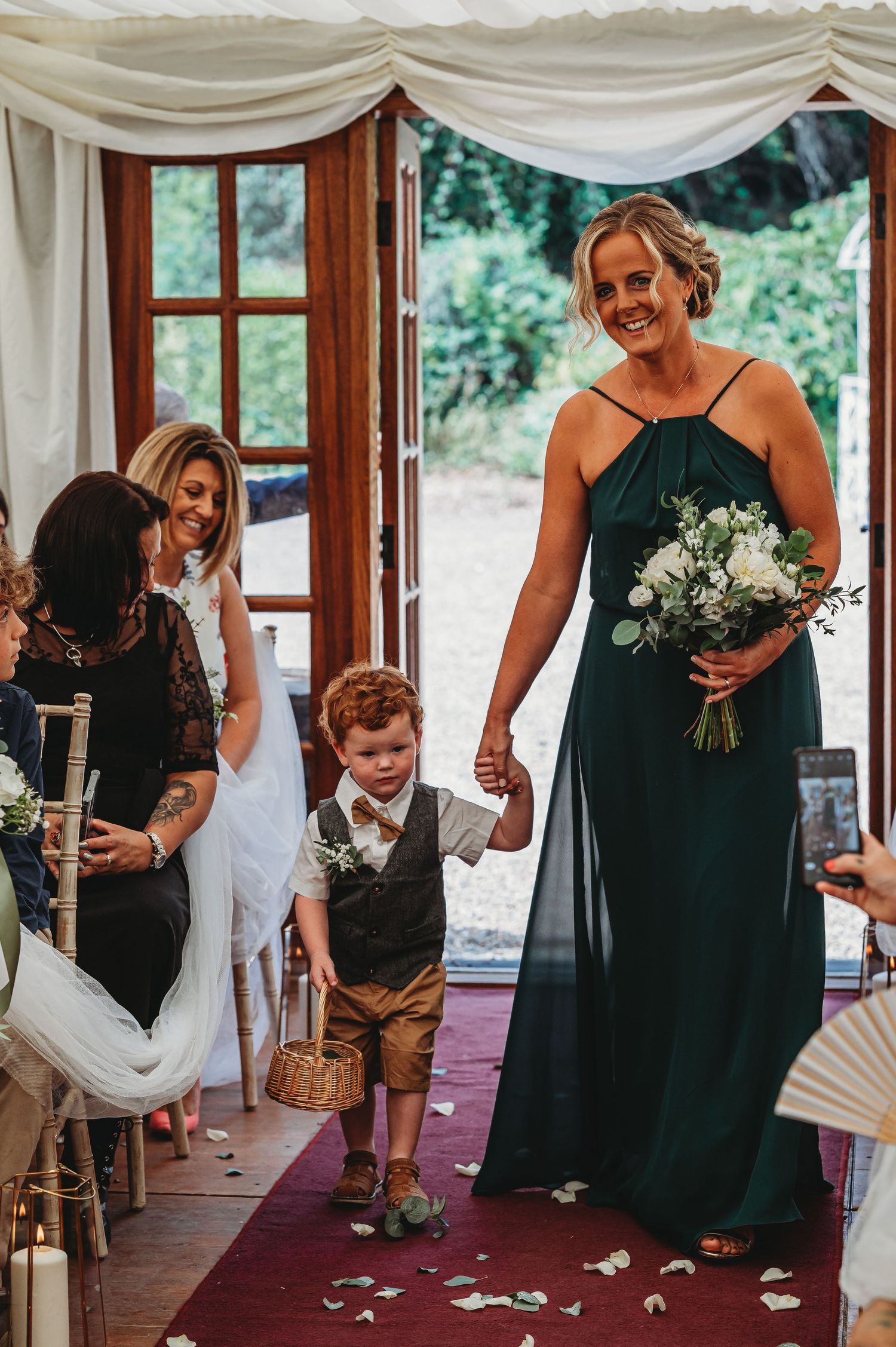 Wedding scene showing ceremony entrance with attendants in dark green dress and formal attire walking down red carpet aisle.