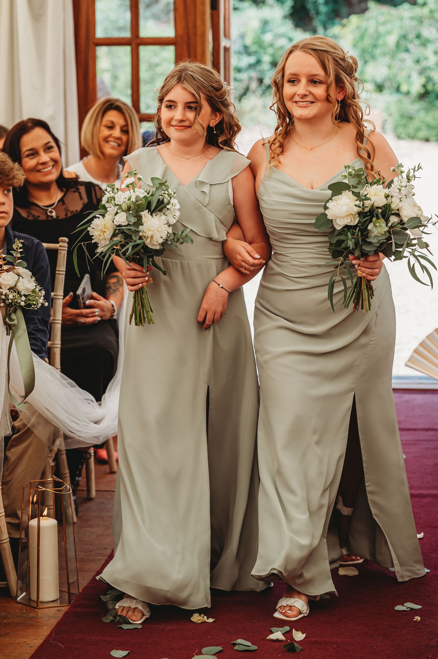Bridesmaids walking arm in arm wearing elegant sage green dresses and holding white floral bouquets during wedding ceremony.