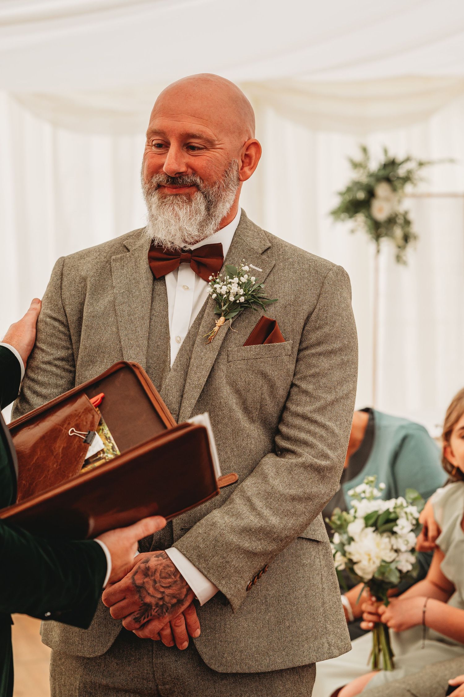 Person in tweed suit and bow tie smiling at wedding ceremony with a small bouquet boutonniere.