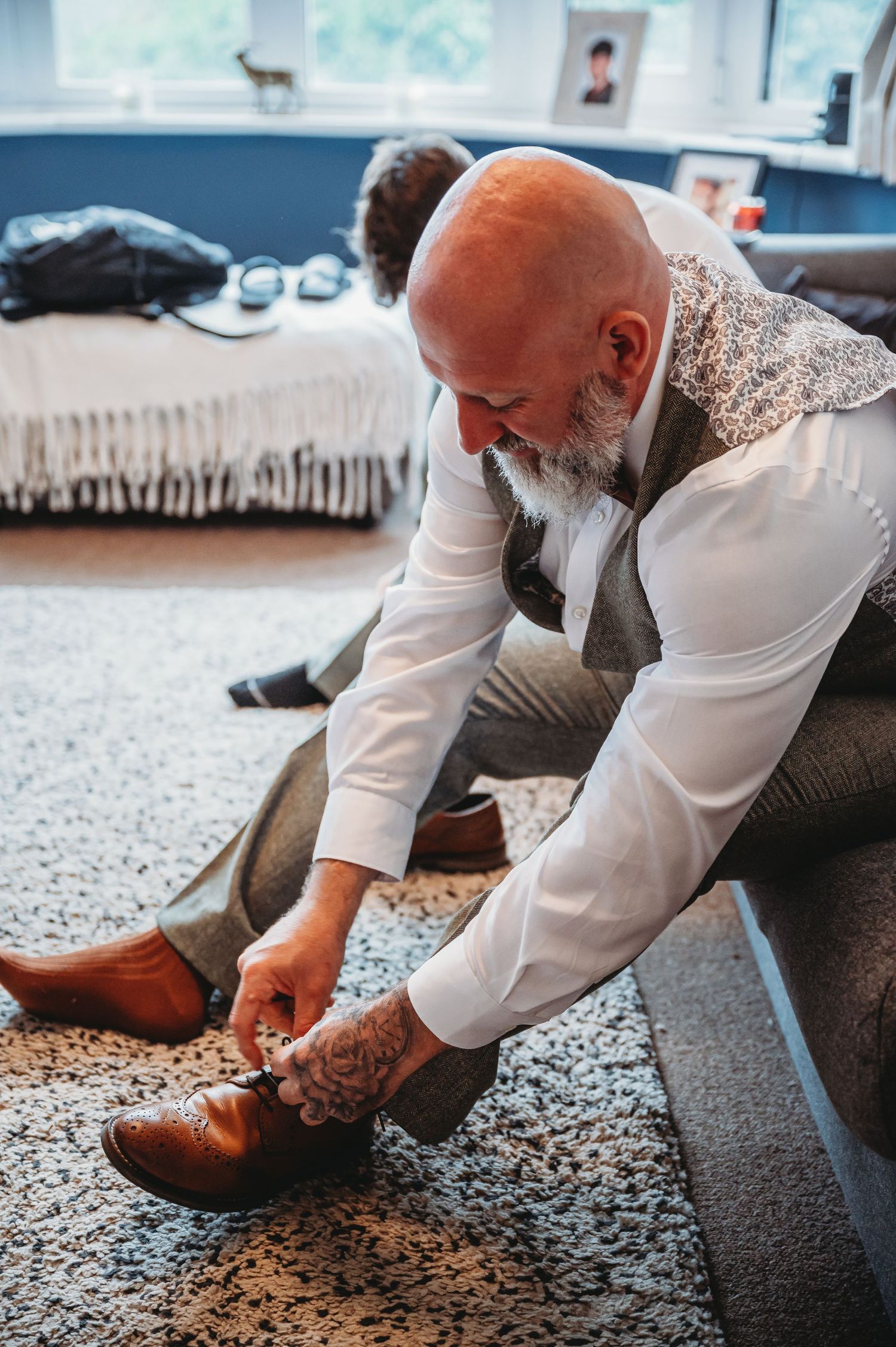 A bearded man wearing a white shirt and vest sits on the floor to put on brown leather dress shoes.