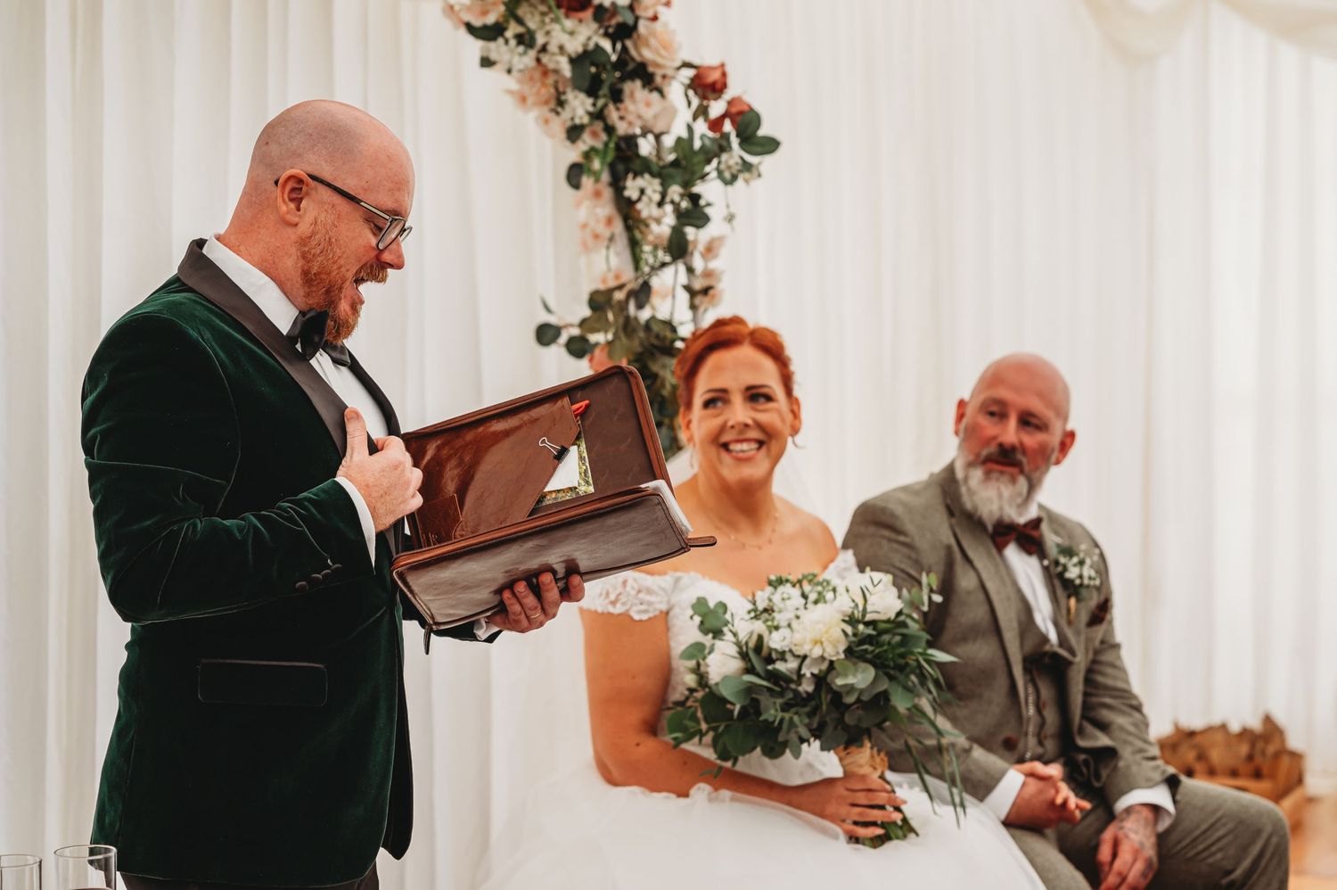 A wedding ceremony moment with three people smiling during a special reading from a leather-bound book.