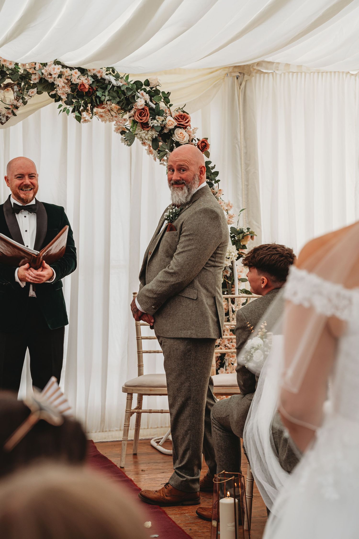 Inside a white wedding tent, people gather under a floral arch during an intimate ceremony.