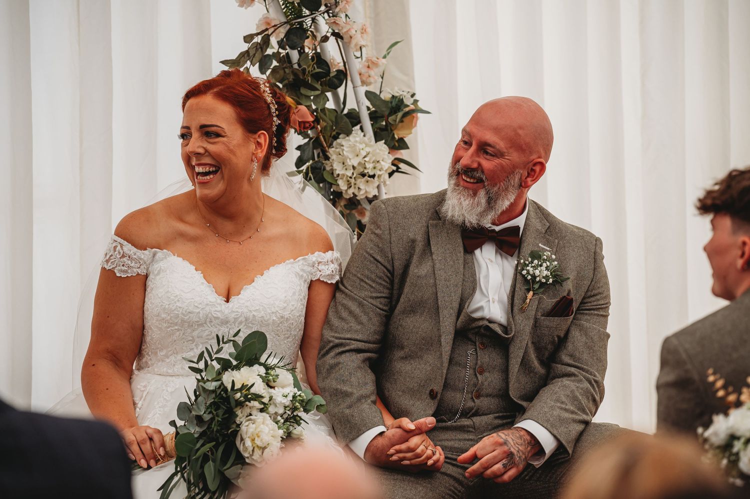 A couple shares a joyful moment during their wedding ceremony in front of white floral decorations.