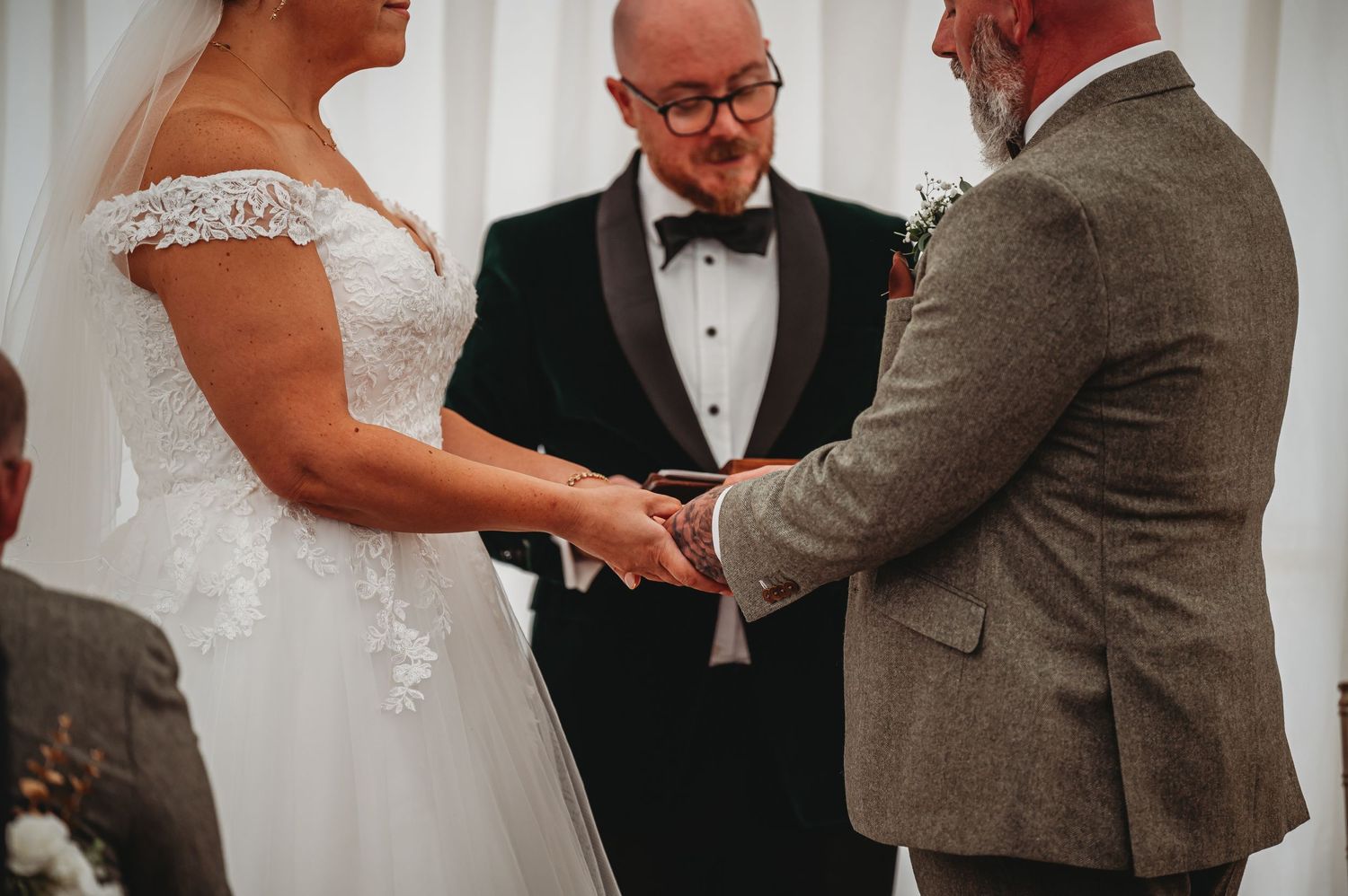 A wedding ceremony taking place indoors with the couple exchanging rings as an officiant in a tuxedo presides.