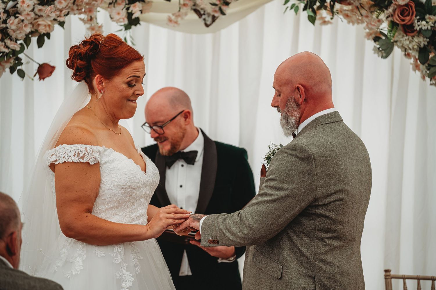 A couple exchanges wedding rings during an indoor ceremony beneath a floral arch with a white backdrop.