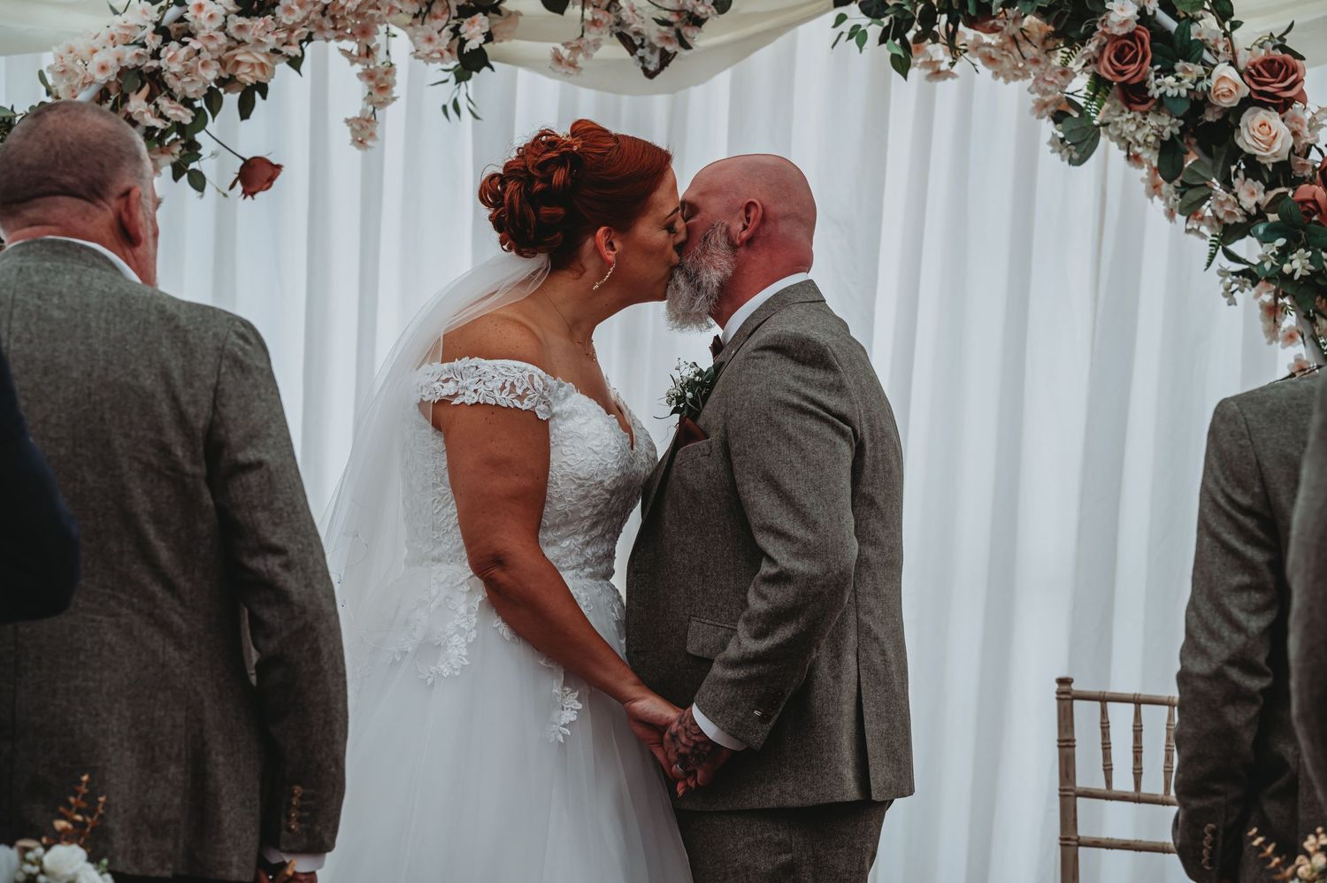 Couple shares intimate moment during wedding ceremony under floral arch with white backdrop and elegant decor.